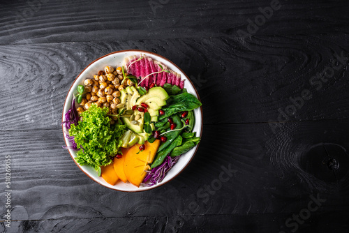 Fresh salad with roasted chickpeas, avocado, persimmon, spinach, avocado, watermelon radish and seeds on a dark background. Long banner format. top view