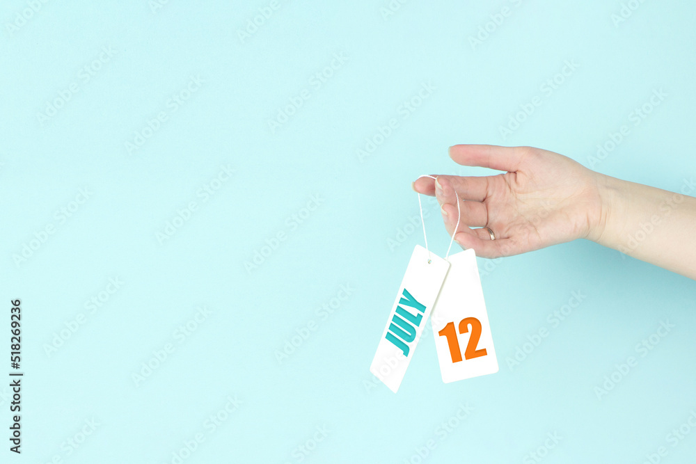 July 12nd. Day 12 of month, Calendar date. Hand hold price tag with calendar date on blue background. Summer month, day of the year concept. July 12nd. Day 12 of month, Calendar date. Hand hold price tag with calendar date on blue background. Summer month, day of the year concept.