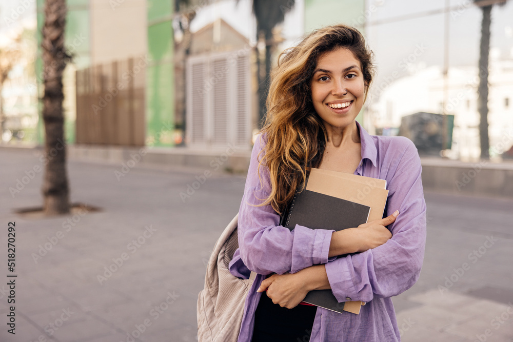 Cute young caucasian girl looks at camera, holds notebooks resting on street after class. Brown-haired female student wears casual clothes. Leisure concept