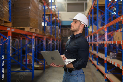 Young caucasian male worker wearing helmet using laptop in modern warehouse.