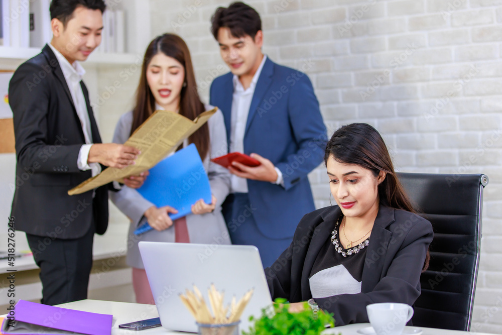 Asian professional successful businesswoman employee sitting working with laptop notebook computer on workstation desk while male and female colleagues standing talking discussing together in office Asian professional successful businesswoman employee sitting working with laptop notebook computer on workstation desk while male and female colleagues standing talking discussing together in office