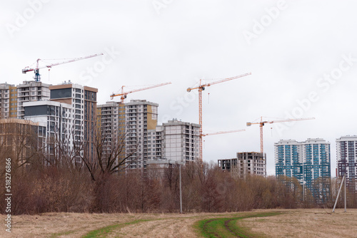Tall residential buildings under construction in the far away. A dirt road is leading to the side of the frame. Several cranes on the background