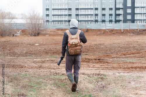A young man with a hunting backpack is walking near a high building on the cold autumn weather holding a long rod. Back view