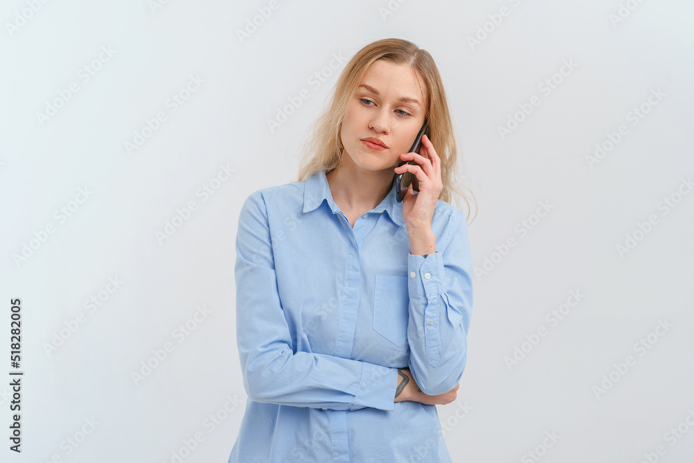Upset and unhappy blonde woman talking on mobile phone, conversation on smartphone, standing over white background in casual blue shirt. Cellphone, cellular concept Upset and unhappy blonde woman talking on mobile phone, conversation on smartphone, standing over white background in casual blue shirt. Cellphone, cellular concept