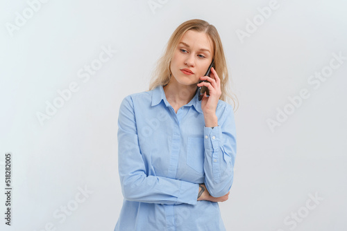 Upset and unhappy blonde woman talking on mobile phone, conversation on smartphone, standing over white background in casual blue shirt. Cellphone, cellular concept