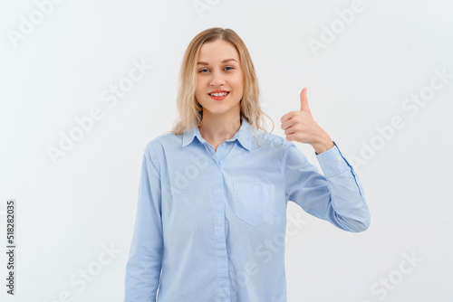 Happy good-looking young blonde woman in in blue casual style shirt, laughing and doing positive gesture with hand, thumb up. Indoor studio shot on white background