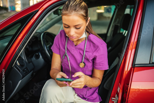 A smiling young nurse dressed in her scrubs uniform sitting in her car holding her mobile phone, taking a break from work. photo