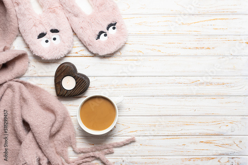 Cozy autumn and winter composition with cup of hot coffee with milk, candle, funny fluffy slippers and warm blanket on white wooden background top view, copy space for your design.