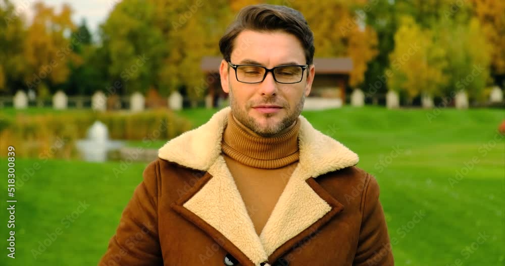 Young handsome man with beard wearing casual sweater and glasses doing happy thumbs up gesture with hand. a man in a jacket and sweater and glasses looks at the camera in the park and shows ok Young handsome man with beard wearing casual sweater and glasses doing happy thumbs up gesture with hand. a man in a jacket and sweater and glasses looks at the camera in the park and shows ok
