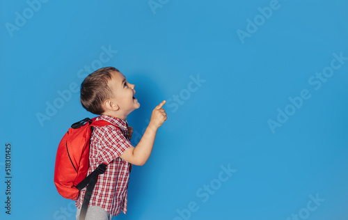 A cheerful smiling little brunette boy with a backpack is having fun on the background of a blue wall and points to your text. The concept of the school. back to school