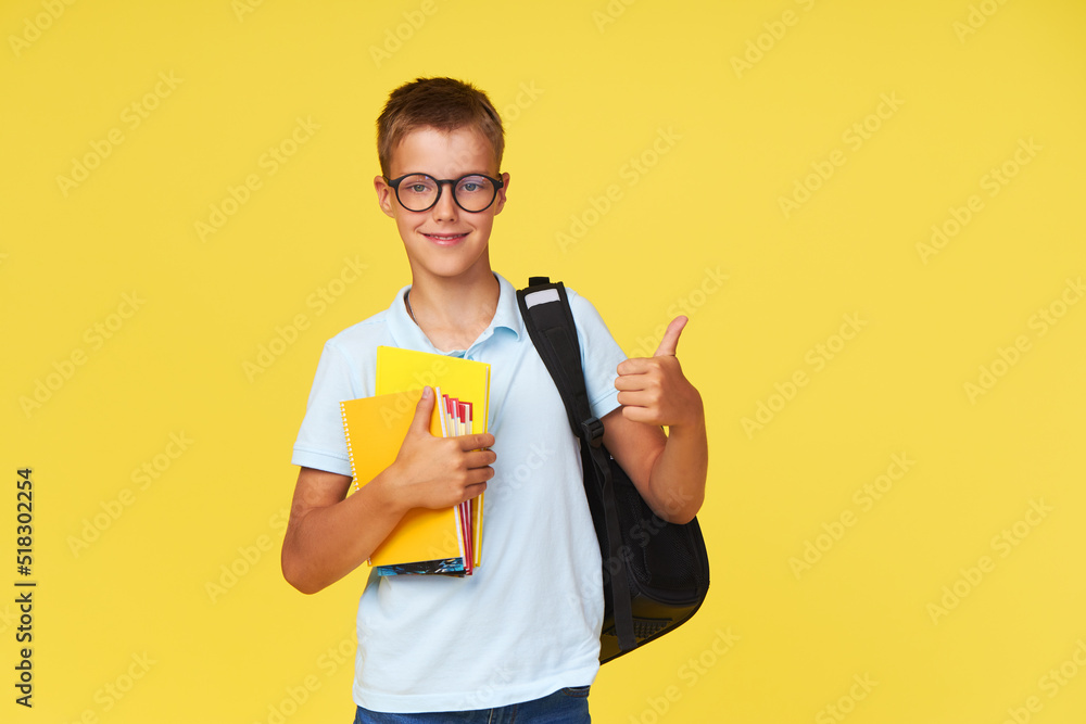 Portrait of a schoolboy in glasses with textbooks and a backpack on a yellow background showing ok sign. Back to school Portrait of a schoolboy in glasses with textbooks and a backpack on a yellow background showing ok sign. Back to school