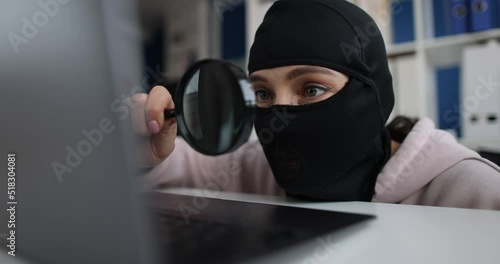 Woman hacker in a balaclava with a magnifier near a laptop photo
