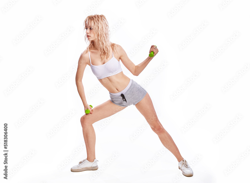 Fit young blonde woman holds green dumbbells isolated on a white background. A sporty slender woman with flat abs does exercises dressed in crop top bra and shorts isolated on white background