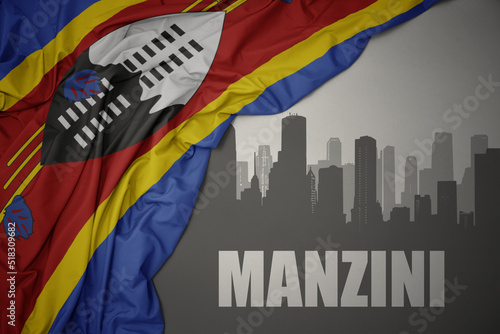 abstract silhouette of the city with text manzini near waving colorful national flag of eswatini on a gray background. photo