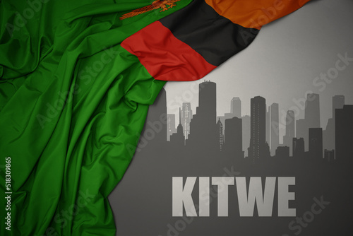 abstract silhouette of the city with text Kitwe near waving colorful national flag of zambia on a gray background.