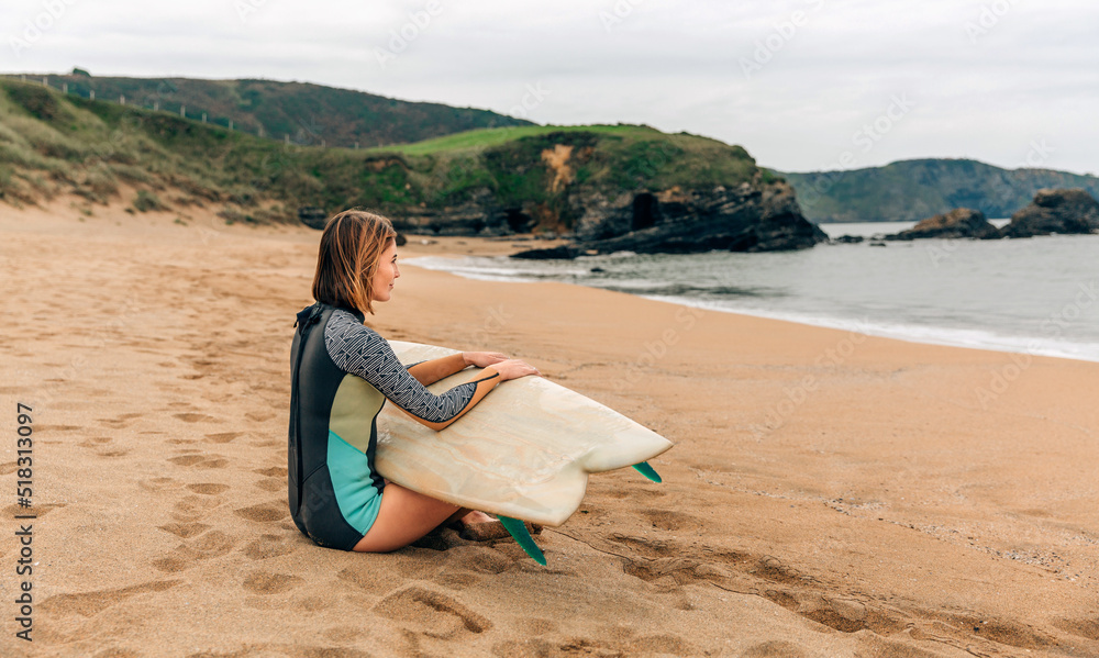 Young surfer woman with wetsuit and surfboard sitting on the sand looking at the sea
