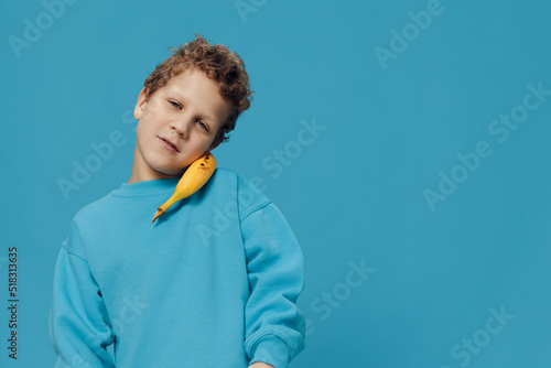 joyful, happy, curly boy stands with a banana near his ear on a blue background in a blue sweater and smiles looking at the camera. Horizontal photo with empty space for advertising mockup