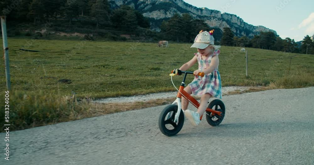 Happy carefree toddler girl cycling on balance bike at empty mountain road along field on evening. Children smile and ride on cycle at summer vacation. Cheerful childhood of preschool girl on country Happy carefree toddler girl cycling on balance bike at empty mountain road along field on evening. Children smile and ride on cycle at summer vacation. Cheerful childhood of preschool girl on country