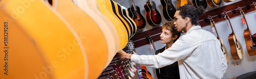 African american seller and customer looking at blurred acoustic guitars in music store, banner.