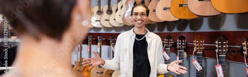 African american seller pointing with hands near acoustic guitars and blurred customer in music store, banner.