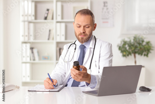 Young male doctor sitting in an office holding a bottle of pills and writing