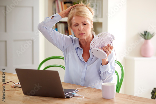 Menopausal Mature Woman Having Hot Flush At Home Cooling Herself With Fan Connected To Laptop photo