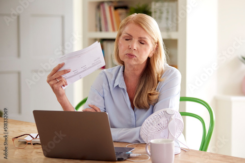 Menopausal Mature Woman Having Hot Flush At Home Cooling Herself With Fan Connected To Laptop photo