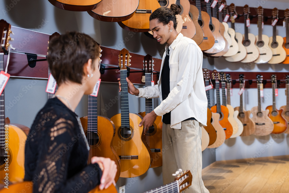 Positive african american seller holding acoustic guitar near blurred  customer in instrumental music store. Photos | Adobe Stock