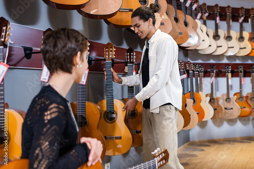 Positive african american seller holding acoustic guitar near blurred customer in instrumental music store.