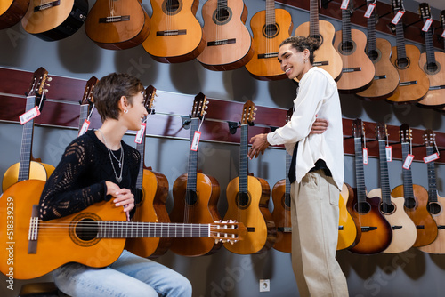 Smiling woman holding acoustic guitar near african american seller in instrumental music store.