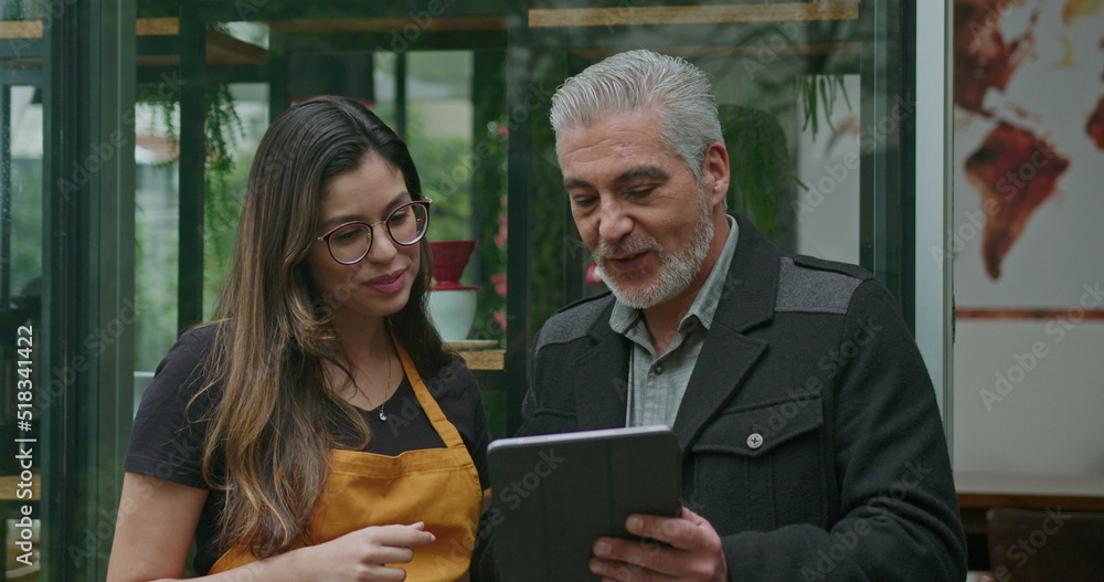 © Marco - Small business owner speaking with female employee barista wearing apron. Boss manager talking with staff explaining restaurant business holding tablet device