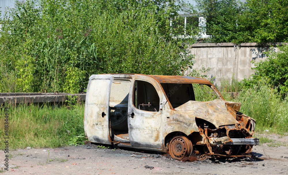 The skeleton of a burnt-out car near a concrete fence overgrown with greenery, Bolshaya Yablonovka Street, St. Petersburg, Russia, July 2022