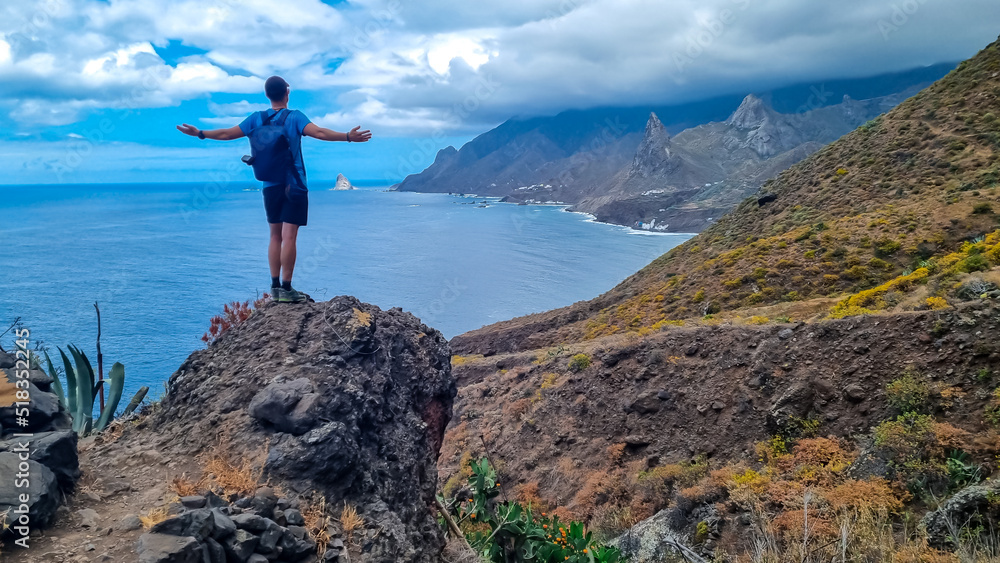 Backpack man with scenic view of Atlantic Ocean coastline and Anaga mountain range on Tenerife, Canary Islands, Spain, Europe. Looking at Roque de las Animas crag. Hiking trail from Afur to Taganana