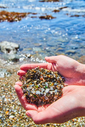 Two hands holding a handful of beach sand filled with shiny and colorful pieces of smooth glass