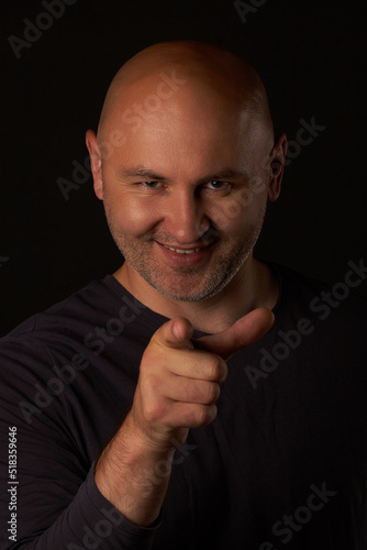 Laughing bald man on a black background points his finger at the camera, vertical photo. A smiling, sly man points his index finger at the camera. You are being deceived. © Aleksandr