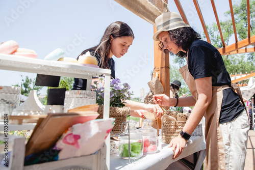 Young brunette girl buyer with long hair and woman seller in apron and hat on either side of the counter with goods at the street fair in summer