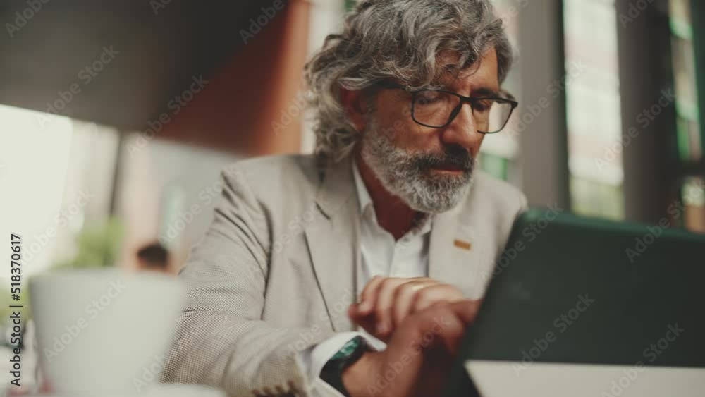 Mature businessman with beard in eyeglasses wearing gray jacket, working on tablet, sitting in an outdoor cafe. Successful man sitting at table in cafe in nature Mobile office freelancer Mature businessman with beard in eyeglasses wearing gray jacket, working on tablet, sitting in an outdoor cafe. Successful man sitting at table in cafe in nature Mobile office freelancer