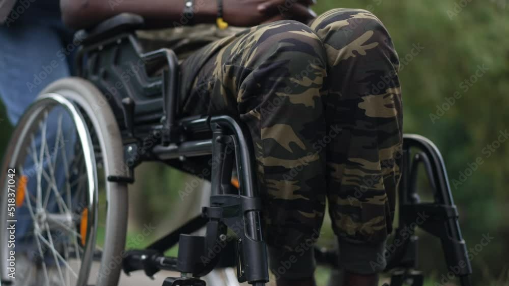 Legs of unrecognizable African American soldier in wheelchair with Caucasian woman pushing mobility aid device outdoors. Young brave ill man supported by girlfriend taking care of partner Legs of unrecognizable African American soldier in wheelchair with Caucasian woman pushing mobility aid device outdoors. Young brave ill man supported by girlfriend taking care of partner