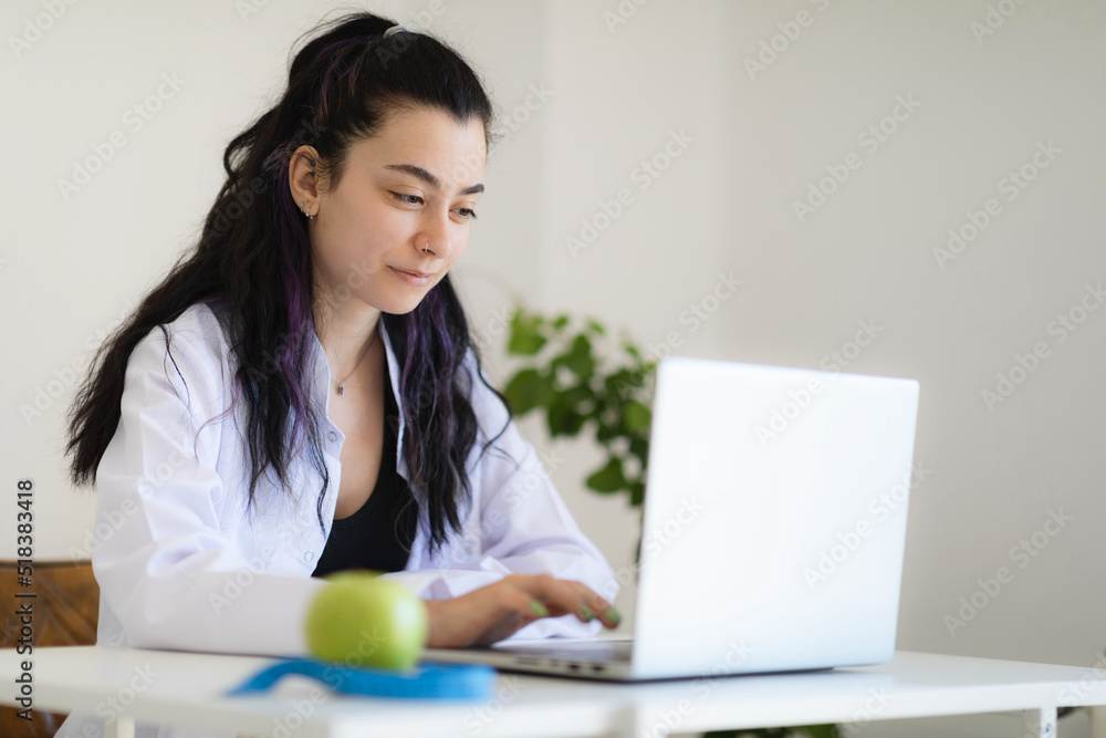 Nutritionist doctor sitting at her cabinet in weight loss clinic, using laptop, giving online consultation about healthy eating and meal plan program. Nutritionist doctor sitting at her cabinet in weight loss clinic, using laptop, giving online consultation about healthy eating and meal plan program.