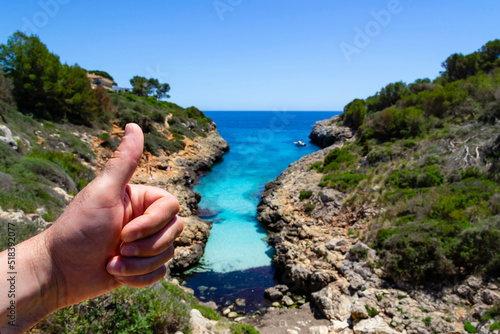 Close up of hand showing thumb up with beautiful blurred seascape in the background with turquoise-blue water in a narrow bay with stony cliffy shore covered with green plants. Active vacation at sea photo