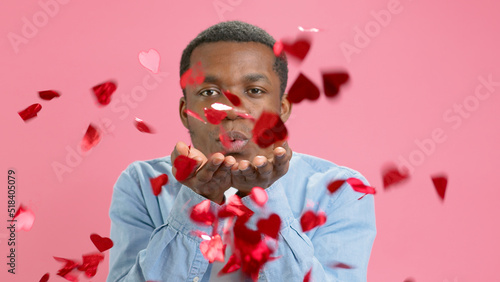 Portrait of carefree African American cheerful teenage man in blue shirt blowing red heart shaped confetti, enjoying birthday or valentines day, festive mood. Indoor studio shot on pink background.