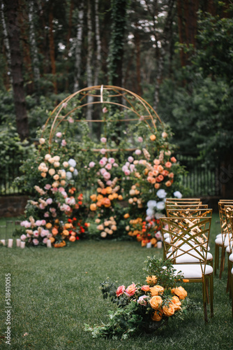 Fototapeta Naklejka Na Ścianę i Meble -  Golden chairs stand in a row on the lawn at an outdoor ceremony in the forest. In the background is a wedding arch decorated with roses and tall pines.