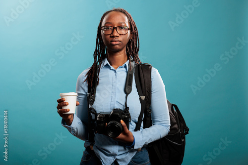 Serious looking professional photographer having DSLR device and cup of coffee while looking at camera on blue background. Young photography entusiast having photo equipment while enjoying beverage. photo