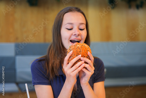 Happy positive smiling teen teenager girl  young woman is holding biting big juicy fat burger  hamburger glass of soda sitting at table on food court. Fast food restaurant  junk unhealthy meal