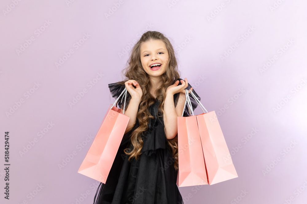 A happy little girl with shopping from a boutique. A child in a black dress holds paper bags on a pink isolated background. A happy little girl with shopping from a boutique. A child in a black dress holds paper bags on a pink isolated background.