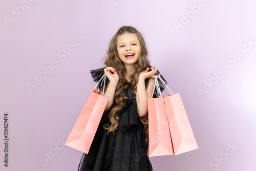 A happy little girl with shopping from a boutique. A child in a black dress holds paper bags on a pink isolated background.