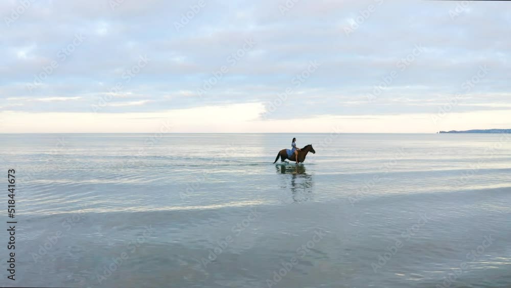 A young beautiful brunette girl with long hair in a blue flowing dress rides a brown horse on Donabate beach, Ireland. A young beautiful brunette girl with long hair in a blue flowing dress rides a brown horse on Donabate beach, Ireland.