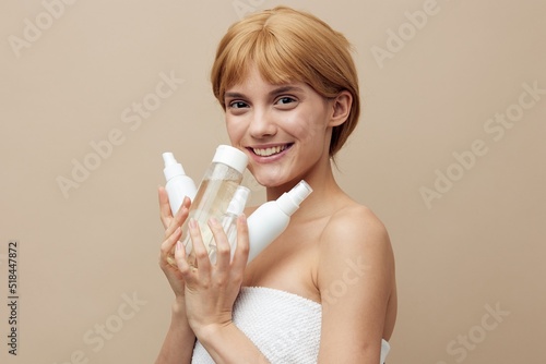 a sweet, young woman stands on a beige background holding various jars of cosmetics in her hands and hides her face from the camera behind them