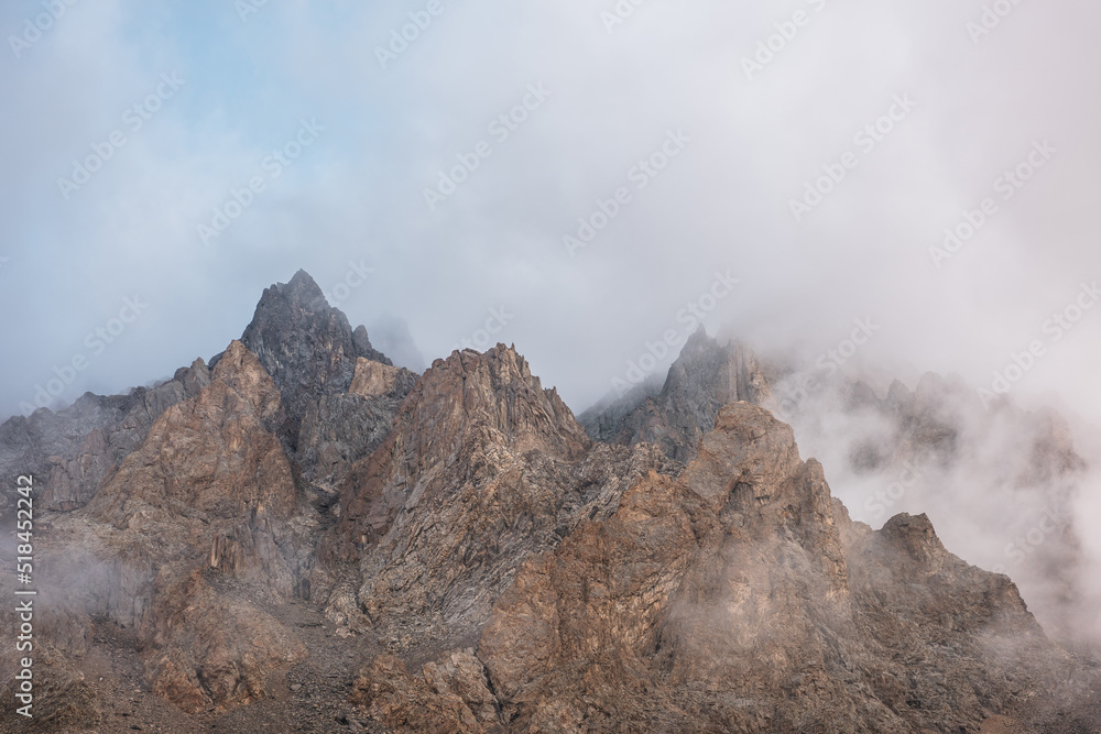 Scenic alpine landscape with rocky mountains in dense low clouds in morning sunlight. Colorful mountain scenery with sharp rocks among thick low clouds. Awesome view to high rockies in low cloudiness.