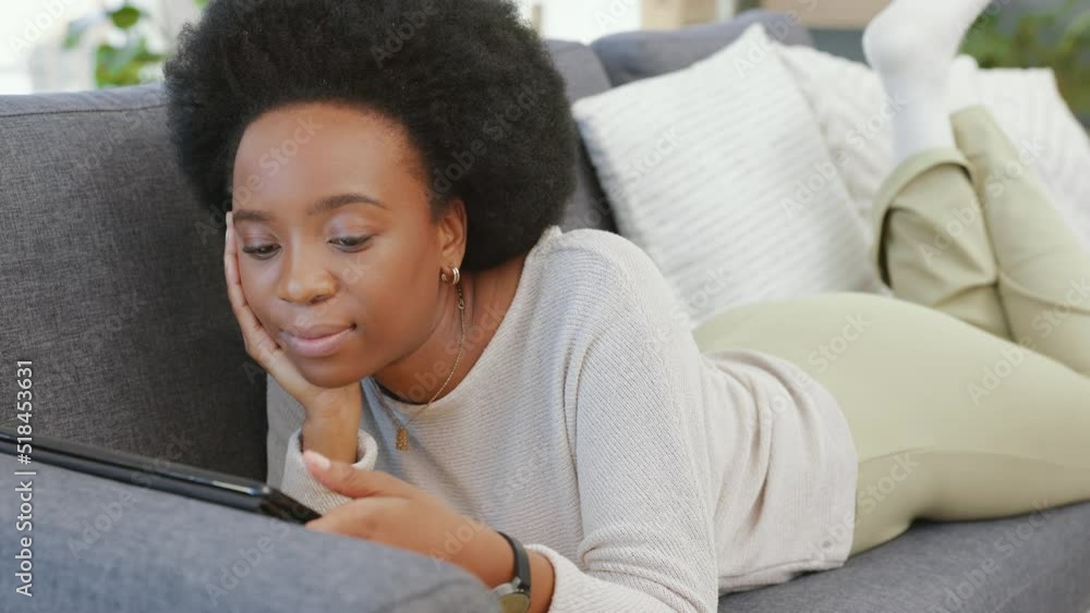 African woman scrolling on a digital tablet while lying on the couch at home. Carefree female with afro hair browsing social media, doing online shopping and enjoying her online movie subscription African woman scrolling on a digital tablet while lying on the couch at home. Carefree female with afro hair browsing social media, doing online shopping and enjoying her online movie subscription